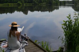 woman reading book while sitting on a wooden dock beside a small body of water relaxing and recharging