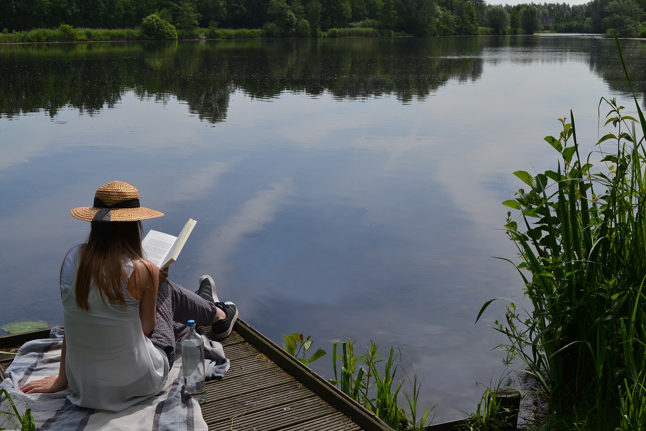 woman reading book while sitting on a wooden dock beside a small body of water