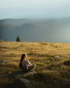 woman sitting on hill cross-legged looking out over valley in peaceful environment