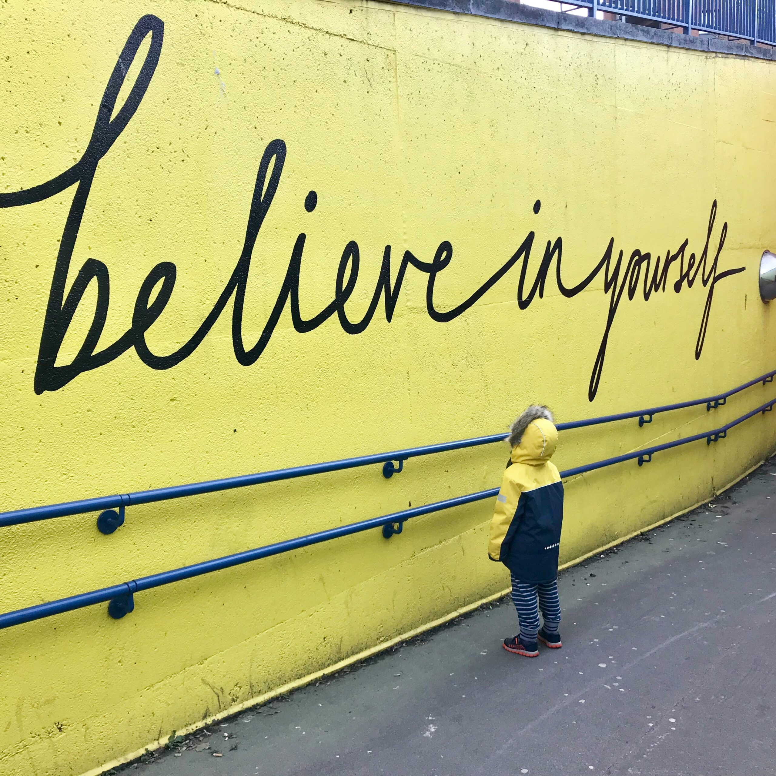 child in yellow and blue rain coat looking a yellow-painted brick wall with scripted writing "believe in yourself"