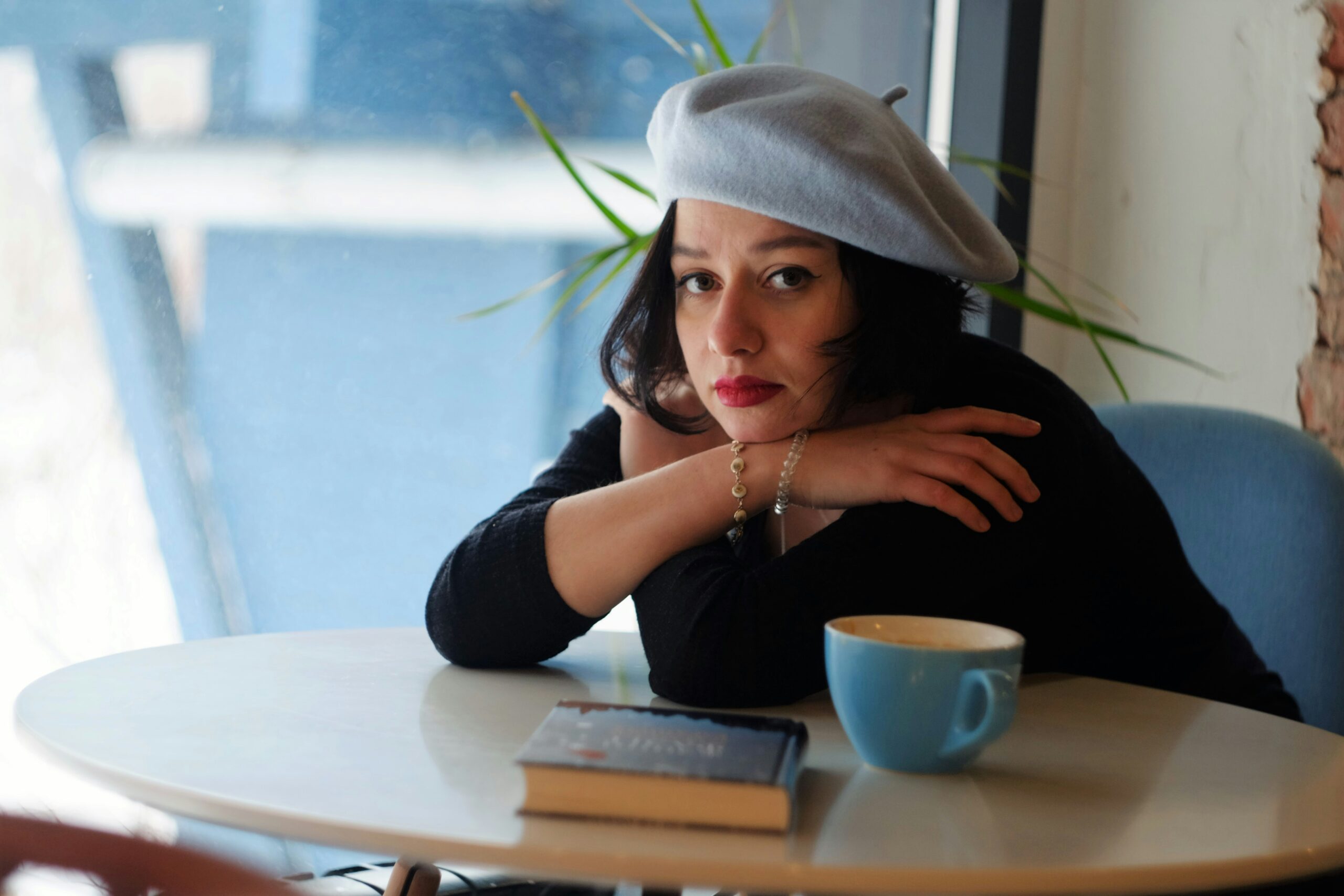 woman with blue beret sitting at cafe table with book and coffee cup leaning on her arms, looking unmotivated