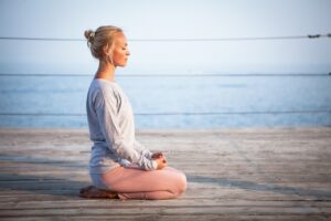 woman sitting on heels on a dock beside a quiet body of water with eyes closed - as if meditating